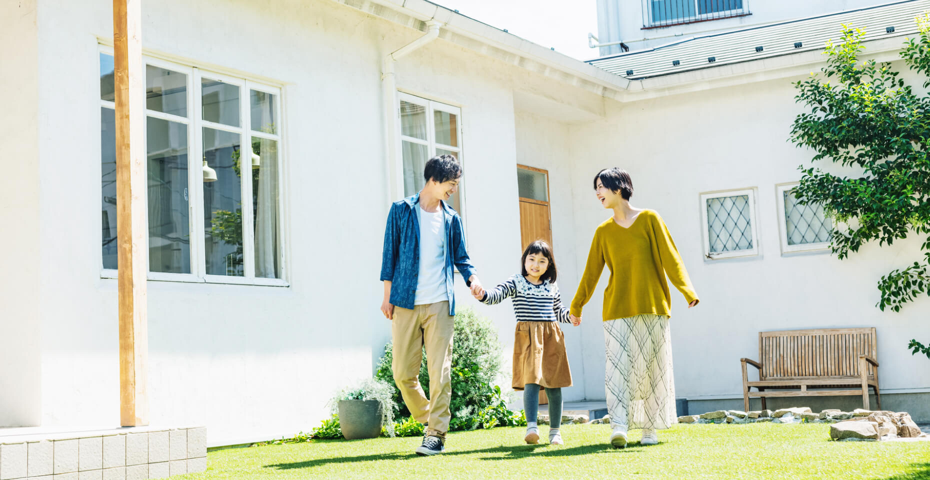 家の前の家族: 白い外壁の住宅の前を、笑顔で散歩する仲睦まじい親子3人。